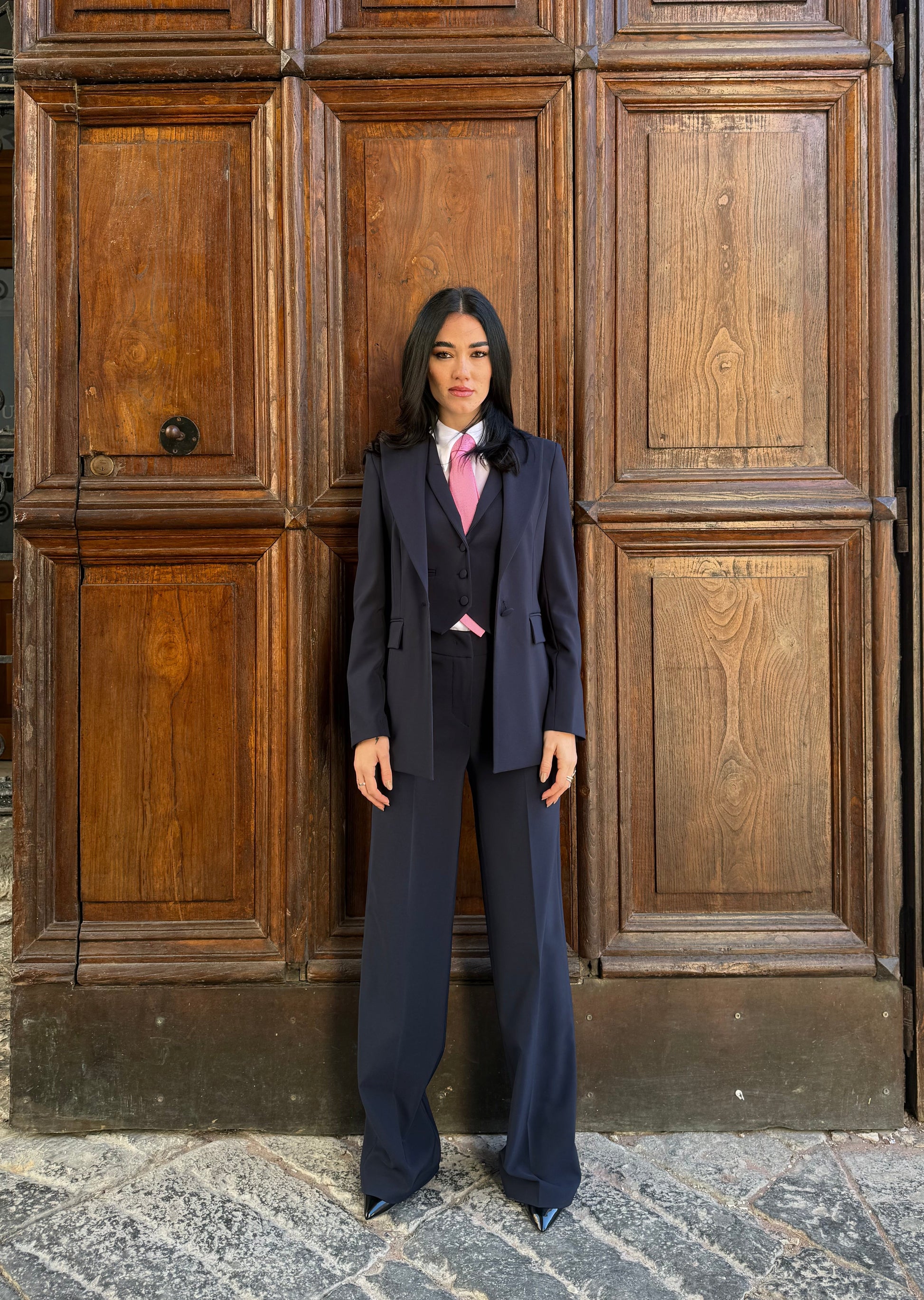 tailleur blu navy con giacca monopetto sfiancata, gilet e pantaloni, donna davanti a portone in legno.